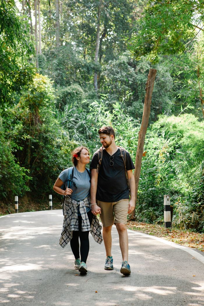 Offering Full body of positive young loving couple holding hands and looking at camera while walking along asphalt road near amidst lush green tropical trees during summer holidays