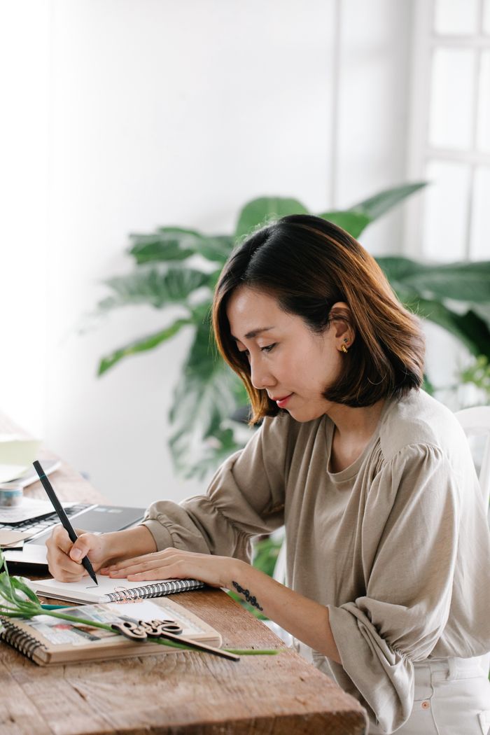 Offering Woman sitting indoors writing in a notebook with greenery in the background, capturing a creative moment.
